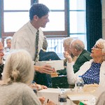 A man holding a box and paper cup speaks to a woman seated at a table; other people are visible sitting around the table